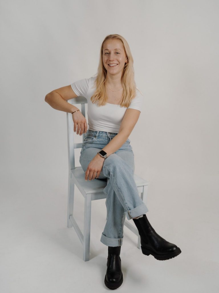 Bild von Luisa Lächelnde Frau in weißem T-Shirt und blauen Jeans sitzt auf einem Stuhl.