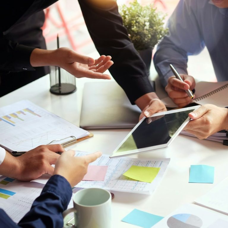 Hände arbeiten an einem Tisch mit Tablets, Notizen und Dokumenten.