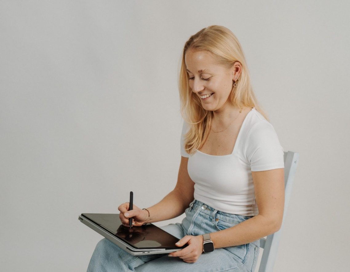 Blonde Frau sitzt lächelnd und arbeitet mit einem Grafiktablett.